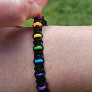 May include: Black macrame bracelet with colorful, rainbow-colored beads. The bracelet features orange, yellow, green, blue, purple, and pink beads. The bracelet is being held by a hand, with a red string visible.