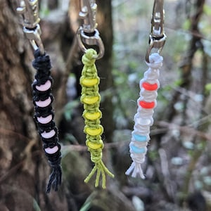 May include: Three handmade keychains hanging from a branch. The keychains are macrame style, with black, white, and pink beads; green and yellow beads; and white, red, and blue beads. Each keychain has a silver clasp.