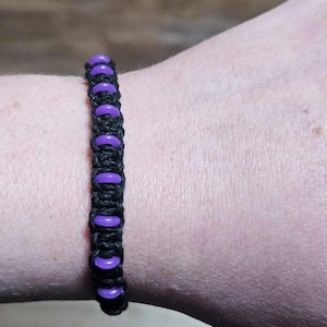May include: A black macrame bracelet with purple beads. The bracelet is worn on a wrist, showcasing the contrast between the dark cord and the vibrant purple accents. The beads are evenly spaced along the length of the bracelet.