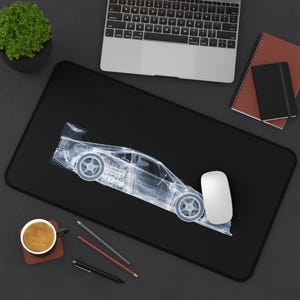 Peut inclure: Tapis de souris rectangulaire noir avec une image transparente et stylisée d'une voiture de course. Une souris d'ordinateur blanche est à droite de la voiture. Un ordinateur portable, une tasse à café et des articles de papeterie sont également visibles.