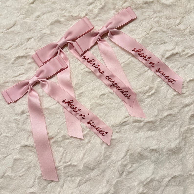 Peut inclure: Trois n&oelig;uds en ruban de satin rose avec le texte "short n sweet" et "sabrina carpenter" brod&eacute; dessus.