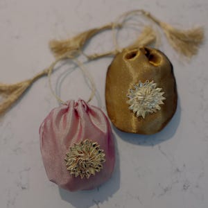 May include: Two small, drawstring pouches. One is a light pink color, and the other is gold. Each pouch has a gold-colored, flower-shaped embellishment on the front. The pouches have matching gold-colored drawstrings with tassels.