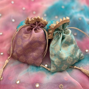 Puede incluir: Dos pequeños y ornamentados bolsos con cordón. Uno es malva, el otro es verde azulado, ambos con estampados florales dorados. Cada bolso tiene un ribete dorado en la parte superior y una larga borla dorada. Los bolsos están sobre una colorida tela teñida.