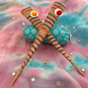 May include: Two decorative henna cones, decorated with colourful threads and small pom-poms, are crossed against a backdrop of patterned fabric in pink, blue, and white. The cones are embellished with gold and various coloured threads.