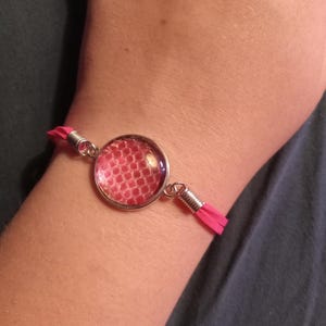 May include: A pink bracelet with a circular, glass-covered charm featuring a red, textured pattern. The charm is connected to pink suede straps with silver-colored metal findings.