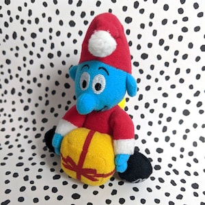 May include: A blue plush toy with a red Santa hat and a yellow gift box. The toy has a white pom-pom on the hat and a white trim around the hat.