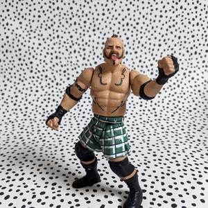 May include: Action figure of a wrestler wearing a green and black kilt, black knee pads, and black wrist bands. The figure has a shaved head, a beard, and tattoos on his chest and arms.