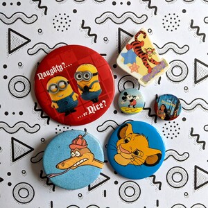 May include: A collection of six colorful round buttons featuring various cartoon characters. The buttons are on a white background with black geometric shapes. The buttons feature characters from Disney and the Minions.