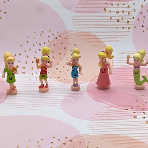 Peut inclure: Cinq poupées en plastique aux cheveux blonds et aux tenues différentes. Les poupées portent une robe verte, une tenue rouge et jaune, une robe bleue, une robe rose et une queue de sirène verte.