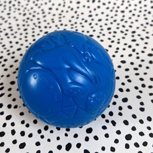 May include: A blue plastic ball with a textured surface and raised designs. The ball is about the size of a baseball.
