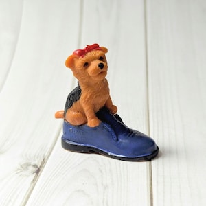 May include: A small figurine of a tan and black dog with a red bow, perched on a blue shoe. The dog has a curious expression. The shoe is a dark blue colour with black trim. The figurine is on a white wooden surface.