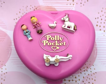 Może przedstawiać: Różowe, sercowate etui Polly Pocket z białym powozem, białym łabędziem, białym koniem i dwiema figurkami. Etui ma napis "Polly Pocket" złotymi literami.