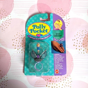 Może przedstawiać: Opakowanie z zabawką Polly Pocket Jewel Surprise Ring. Opakowanie jest turkusowe i fioletowe, z przezroczystym plastikowym okienkiem, które pokazuje małą lalkę i pierścionek. Pierścionek jest fioletowy i srebrny, a lalka ma na sobie niebieski strój. Widoczny jest tekst "Polly Pocket" i "Jewel Surprise Ring".