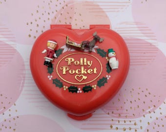 May include: Red heart-shaped Polly Pocket compact with a Christmas theme. It features a small girl doll, a reindeer, a sled, and a snowman. The compact has a gold inscription that reads "Polly Pocket".