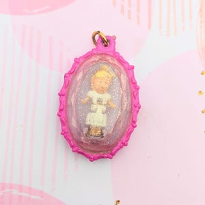 May include: A vintage pink pendant featuring a miniature doll encased in a clear, oval frame. The doll wears a white dress and has blonde hair. The pendant has a gold-coloured loop at the top for hanging.