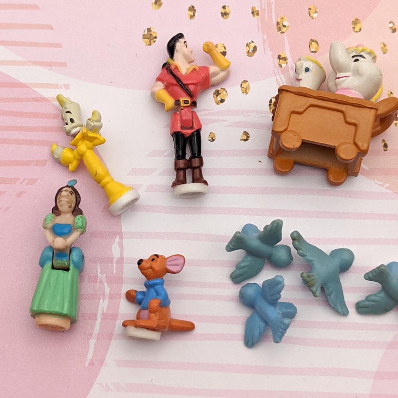 May include: A collection of small plastic figurines featuring characters from Disney movies. The characters include Belle, Beast, Lumiere, Dumbo, and the birds from Peter Pan. The figurines are all in various poses and are painted in bright colors.