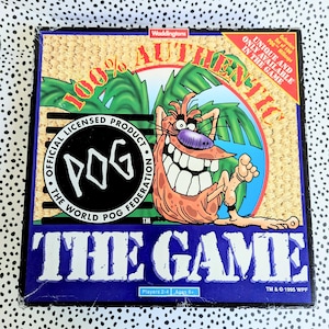 May include: A vintage POG game box with a cartoon character illustration. The box features the text "100% AUTHENTIC" and "THE GAME." The game is an official licensed product of the World POG Federation, for 2-4 players, ages 6+.