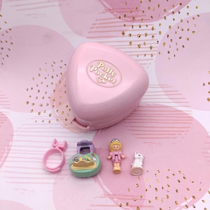 Może przedstawiać: Różowy zestaw do zabawy Polly Pocket z pasującym pierścionkiem, małą lalką, miniaturowym kotem i małym, okrągłym zestawem do zabawy. Zestaw do zabawy jest w kolorze jasnoróżowym i ma nadruk "Polly Pocket". Lalka ma na sobie różową sukienkę.