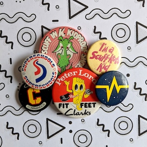 May include: Five colourful button badges with various designs. One badge features a green dragon with the text "Scuff Ink Kidproof Shoes". Another badge has a red and blue logo with the text "Superschools". A third badge has a yellow cartoon foot with the text "Peter Lord Fit Feet Clarks". A fourth badge has a yellow lightning bolt on a blue background. The final badge has the text "I'm a Scuff Kid" in yellow on a red background.