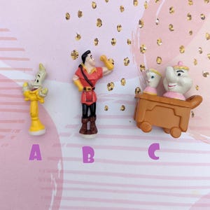 May include: Three small plastic figurines from the Disney movie Beauty and the Beast. The figurines are of Belle, the Beast, and Lumiere. The figurines are yellow, red, and white.