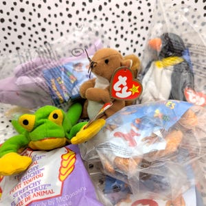 May include: A collection of vintage Beanie Babies in their original packaging. The Beanie Babies include a green frog, a brown squirrel, and a black and white penguin. The packaging is clear plastic with a purple and yellow label that reads "TheTchyy Stretchy Flavestruz" and "Only by McDonald's".