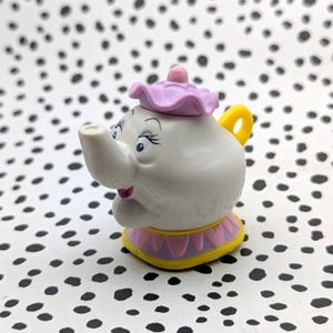 May include: A white teapot with a pink lid and yellow handle. The teapot has a pink and yellow base and a smiling face with blue eyes.