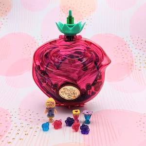 Può includere: Un playset Polly Pocket vintage a forma di rosa rosa. Il playset ha una parte superiore verde e include una piccola bambola e diversi accessori in miniatura. Lo sfondo è un motivo rosa e oro.