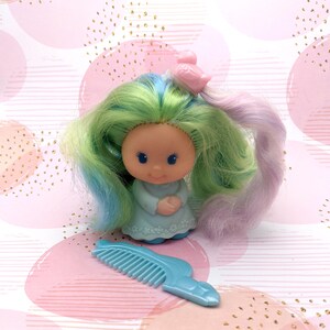 Peut inclure: Une poupée vintage aux cheveux verts et bleus avec une robe bleu clair et un peigne. La poupée a un clip à cheveux en forme d'oiseau dans les cheveux.