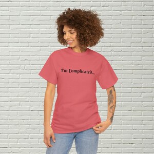 May include: A woman wearing a coral colored t-shirt with the text "I'm Complicated..." printed in black. She is standing in front of a white brick wall.