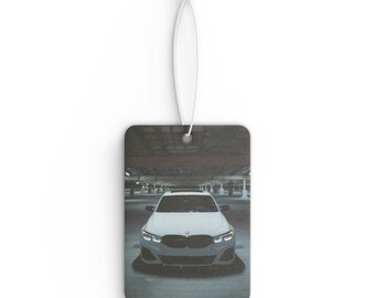 May include: Rectangular car air freshener featuring a white car in a parking garage. The car has its headlights on. The air freshener is attached to a white string.
