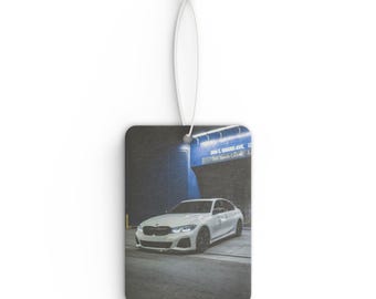 May include: Rectangular car air freshener with a white car parked in a garage. The car is white with black rims. The background is a dark garage with blue and white text.