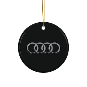 May include: A black, round ornament with the Audi logo in silver. The logo consists of four interlocking rings. A gold string is attached for hanging.
