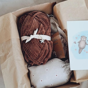 May include: A brown yarn skein tied with a white ribbon and a starfish charm, inside a cardboard box. The box also contains a small, white floral pillow, a card with an otter illustration, and other small items.