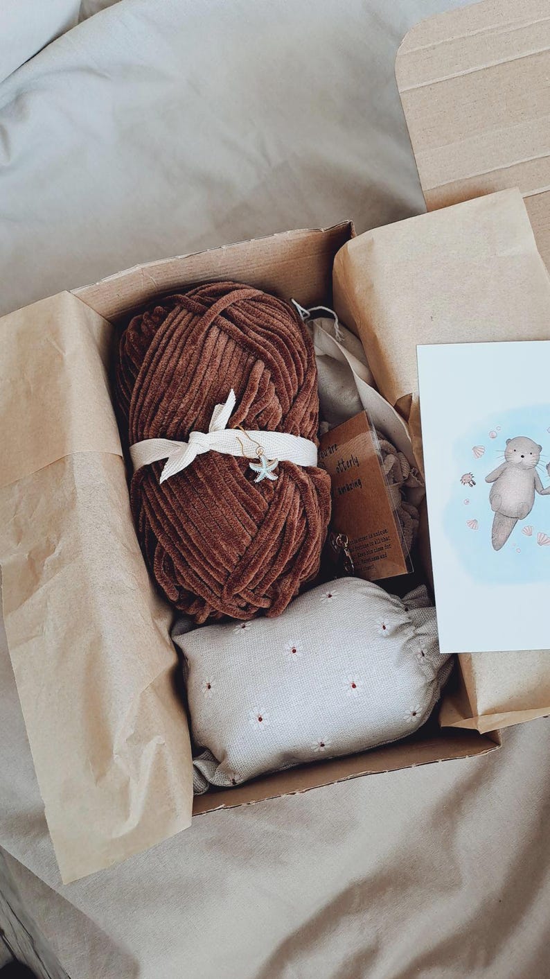 May include: A brown yarn skein tied with a white ribbon and a starfish charm, inside a cardboard box. The box also contains a small, white floral pillow, a card with an otter illustration, and other small items.