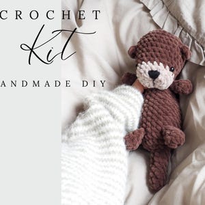 May include: A handmade crochet kit with the text "CROCHET Kit HANDMADE DIY". The kit includes a brown otter plush toy with a cream-colored face and a textured, chunky knit design. The toy is lying on a bed.