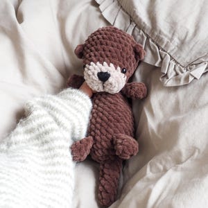 May include: A handmade brown crochet otter plush toy with a cream-colored face and a black heart-shaped nose. The plush toy is held against a neutral-toned background, showcasing its soft texture and detailed craftsmanship. A cozy, handmade item.