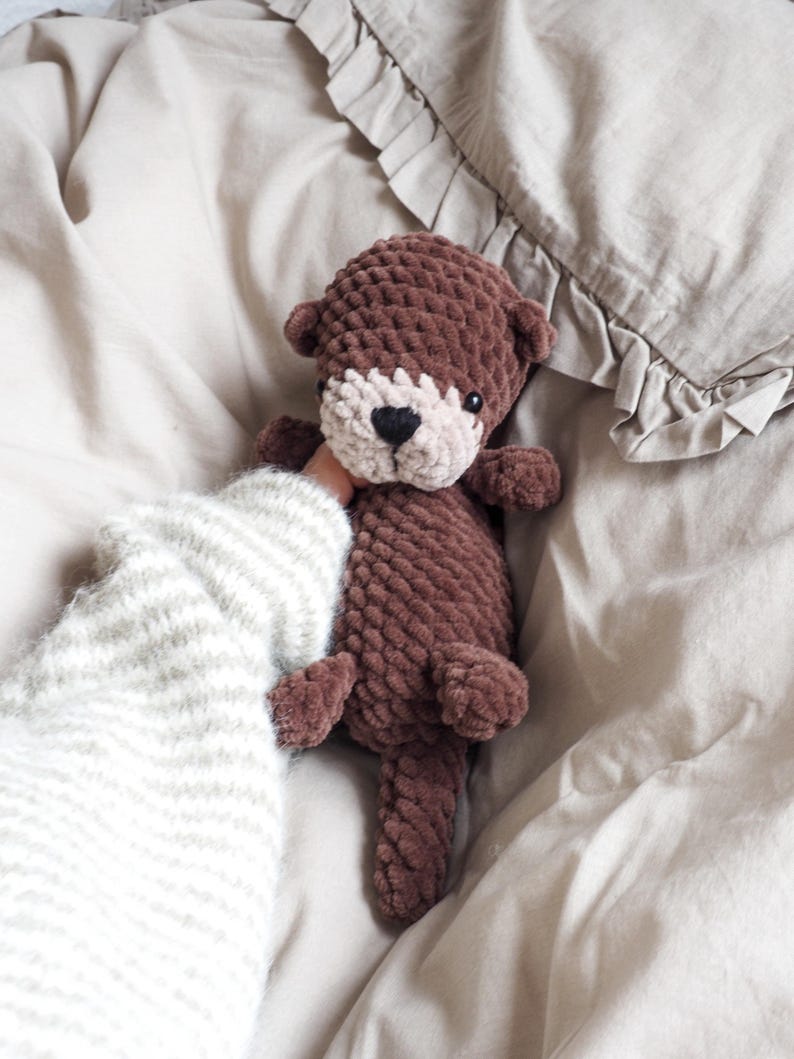 May include: A handmade brown crochet otter plush toy with a cream-colored face and a black heart-shaped nose. The plush toy is held against a neutral-toned background, showcasing its soft texture and detailed craftsmanship. A cozy, handmade item.