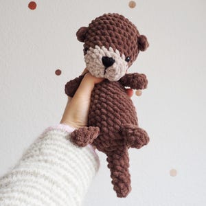 May include: A handmade brown crochet otter plush toy with a light tan face and black nose. The stuffed animal has small ears and is held up against a white background with brown and tan polka dots. The otter's body is a textured, chunky knit.