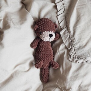 May include: A handmade brown crochet otter plush toy with a white face and black eyes. The plush toy is lying on a beige bedsheet. The otter has small arms and legs and is made of a soft, textured yarn.