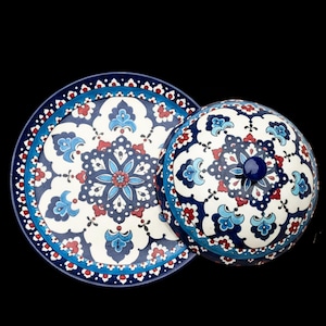 Puede incluir: Un juego de plato y tapa de cerámica con un detallado estampado floral en tonos azules, rojos y blancos. El plato es redondo con una tapa abovedada a juego, ambos sobre un fondo negro. La tapa tiene un pequeño pomo azul.