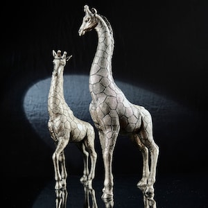 Könnte beinhalten: Zwei silberne Giraffenfiguren, eine größer als die andere, stehen auf einer schwarzen Oberfläche. Die größere Giraffe schaut nach rechts, während die kleinere Giraffe nach links schaut.