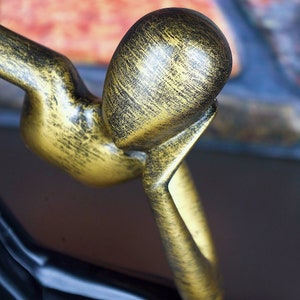 May include: A gold-colored metal sculpture of a person with their head resting on their hand. The sculpture has a distressed finish.