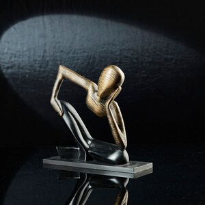May include: A wooden sculpture of a person sitting with their head in their hands. The sculpture is made of dark wood and has a gold finish. It is sitting on a black base.