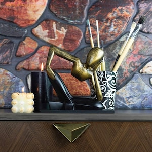 May include: A gold-toned metal sculpture of a person in a seated pose, holding a black and white patterned pencil holder with several pencils inside. Two candles, one black and one white, are in front of the sculpture.
