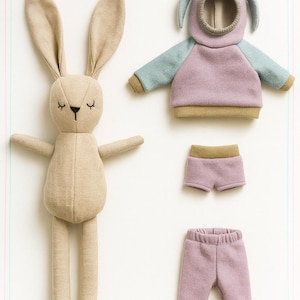 May include: A beige bunny doll with closed eyes and outstretched arms. Beside it are a purple and blue hooded sweatshirt, shorts, and pants. The doll and clothing are made of soft fabric, suggesting a children's toy or craft project.