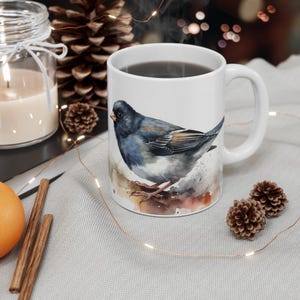 May include: A white ceramic mug featuring a watercolor illustration of a dark-feathered bird with a white belly and orange-brown wing accents. The bird is perched on a branch, depicted with soft, blended paint strokes against a neutral background.