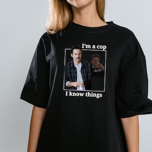 May include: Black t-shirt with a rectangular graphic featuring a man in a plaid shirt and the text "I'm a cop, I know things." The graphic includes a smaller image of the same man.