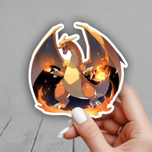 May include: A sticker featuring a Charizard, a fire and flying-type Pokémon. The illustration shows Charizard with orange and yellow scales, black wings, and flames. The sticker is held by a hand with a white painted nail.
