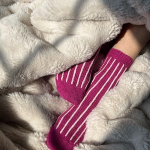May include: A pair of knit socks featuring a vibrant magenta base color with vertical white stripes running along the length of the fabric. The socks have a ribbed texture and are shown extending from beneath a soft, plush, off-white faux fur blanket.