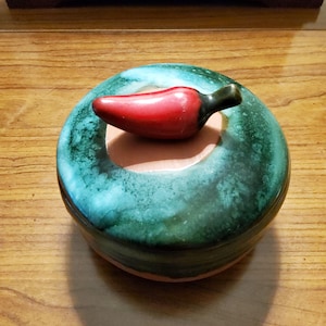 May include: A round ceramic lidded container featuring a glossy, mottled teal and dark green glaze. The lid is topped with a sculpted, smooth red chili pepper handle with a dark brown stem, contrasting against a circular beige center patch.